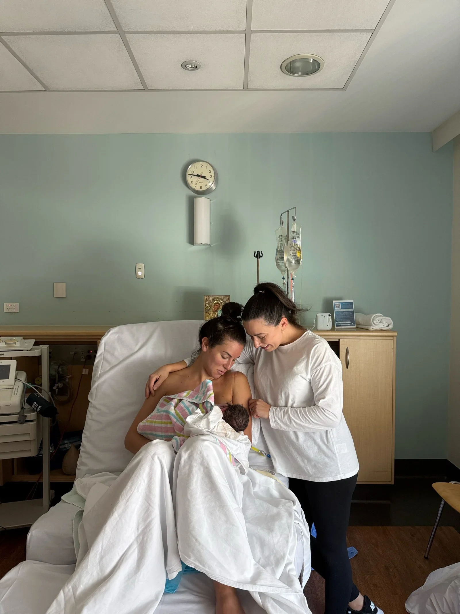 Steph the Doula following the birth of another happy client's baby. Steph looks down and smiles at the mother and baby together in their hospital bed.