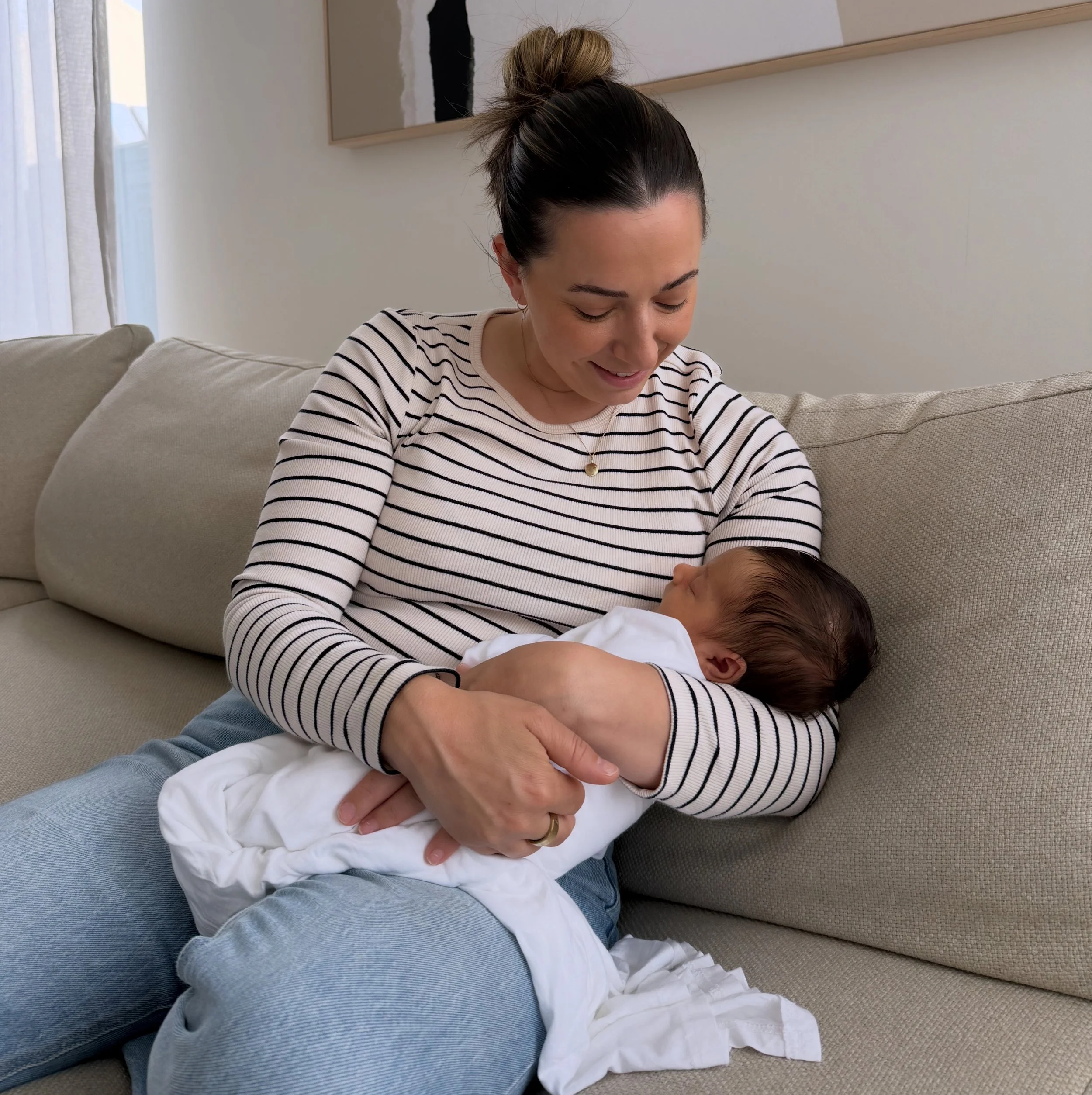 Steph the Doula holding a newborn baby in her client's home while visiting during the early postpartum period. She sits comfortably on the couch with a warm smile on her face, with the baby resting peacefully in her arms.