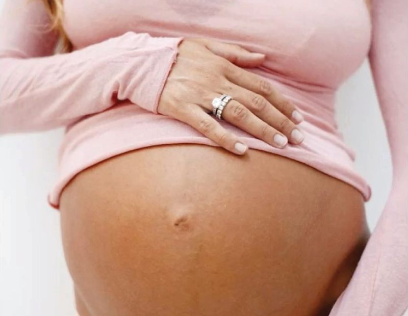 Close-up of a pregnant belly with a hand resting on it, showing rings on the fingers, and a pink shirt pulled up to reveal the belly.