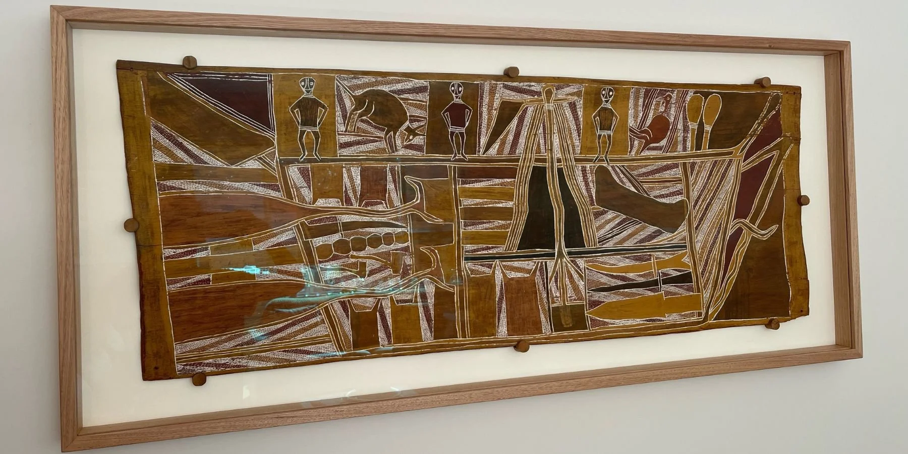 Traditional Aboriginal bark painting mounted in a conservation-standard Tasmanian oak box frame with white backing, featuring figurative and totemic imagery in ochre, brown and white pigments, displayed on a white wall