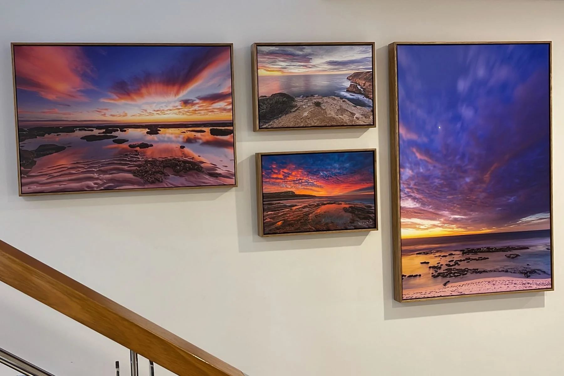Four large landscape photography prints of dramatic coastal sunsets and seascapes in matching gold float frames, installed as a curated wall grouping — two horizontal and two vertical formats arranged asymmetrically on a white wall