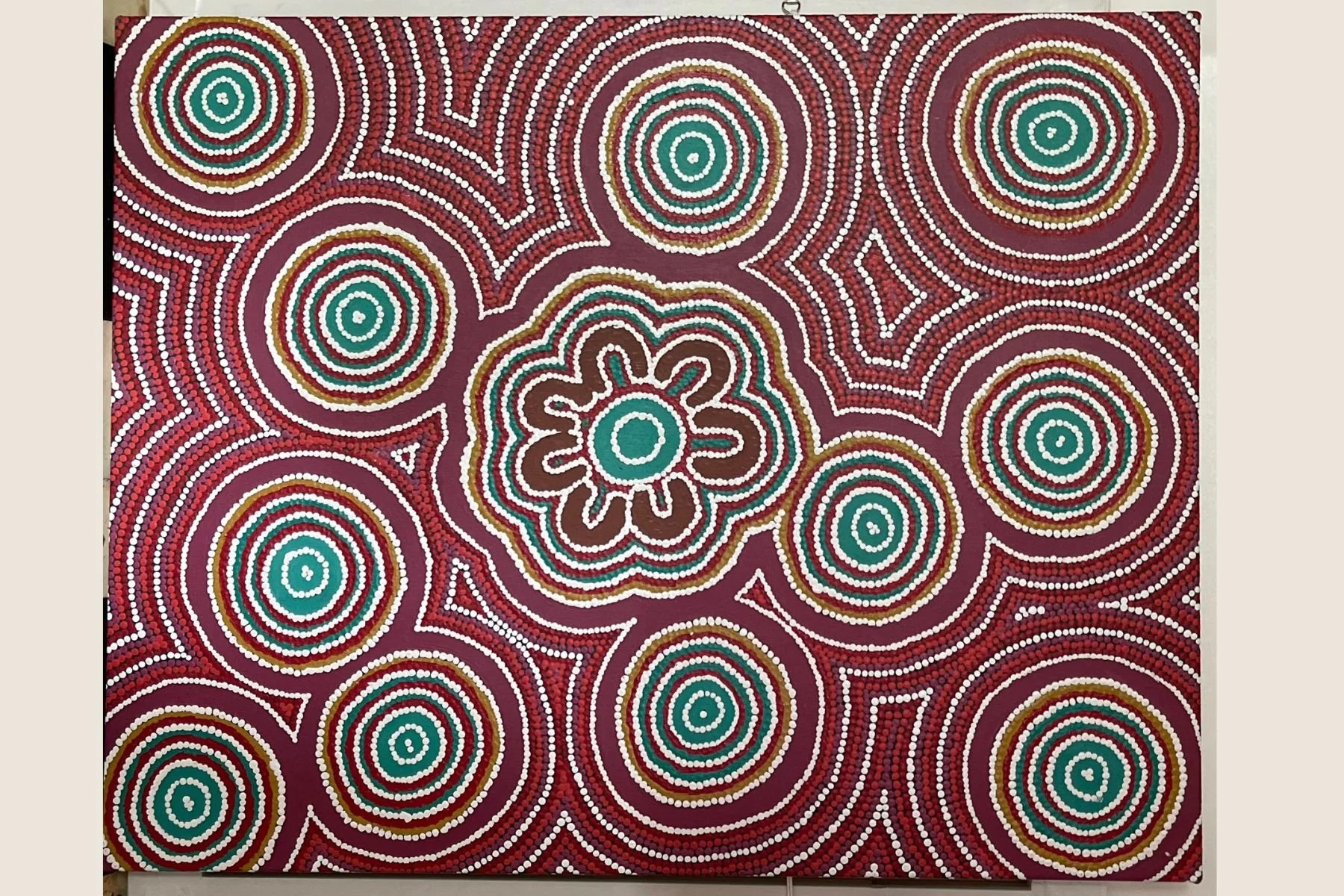 aboriginal dot painting with concentric circles, spirals, and a central flower design in earthy and teal tones on a maroon background.