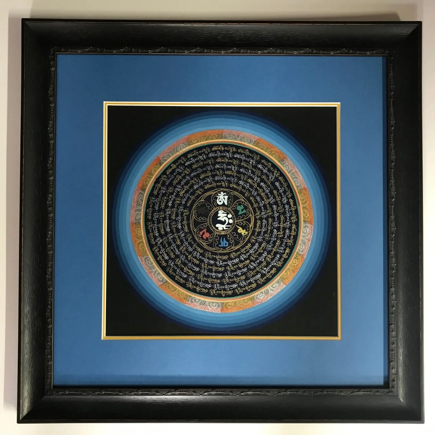 Tibetan Buddhist mandala with concentric rings of gold Sanskrit mantra script on a black ground, with a central Om symbol in a lotus, mounted in a blue and black double mat with a gold fillet inside a black ornate frame