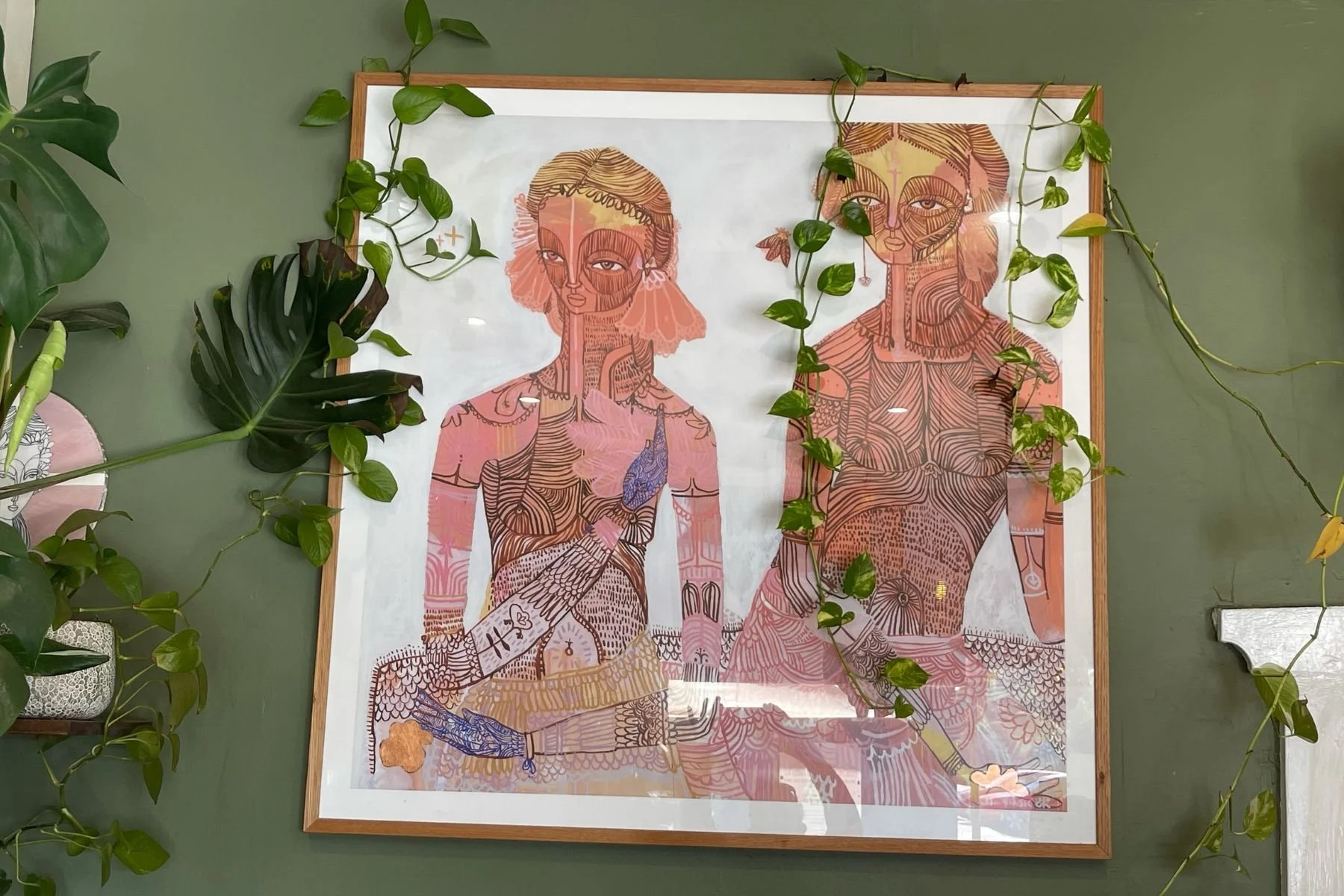 Large contemporary artwork depicting two stylised female figures with intricate linear body markings in warm pinks and ochres, framed in a slim Tasmanian oak frame with white mat, hung on a sage green wall with trailing plants