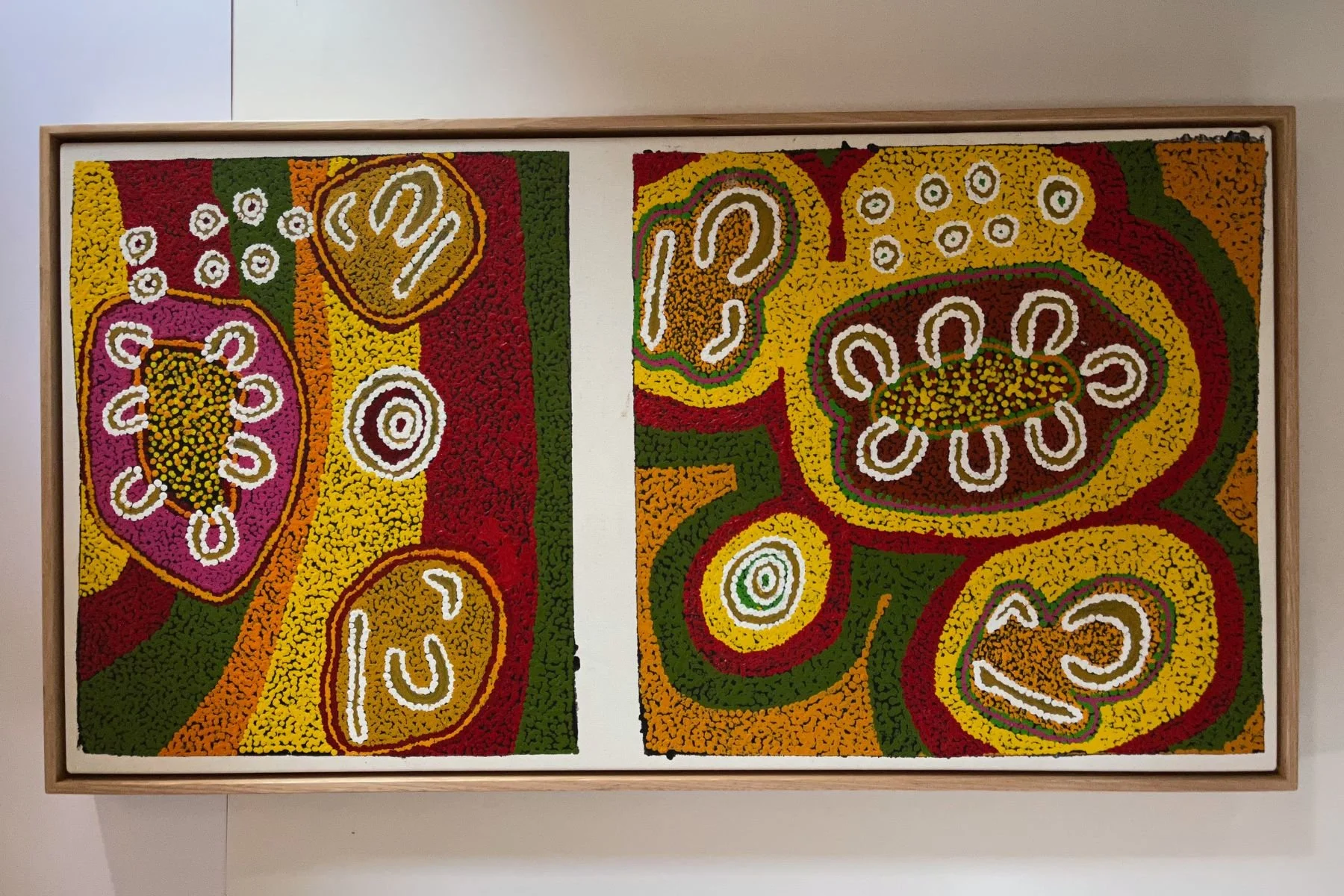 Large Aboriginal diptych of two canvases framed together in a single natural oak float frame, featuring dense dot work in yellows, greens, reds and purples with totemic forms and concentric circle motifs