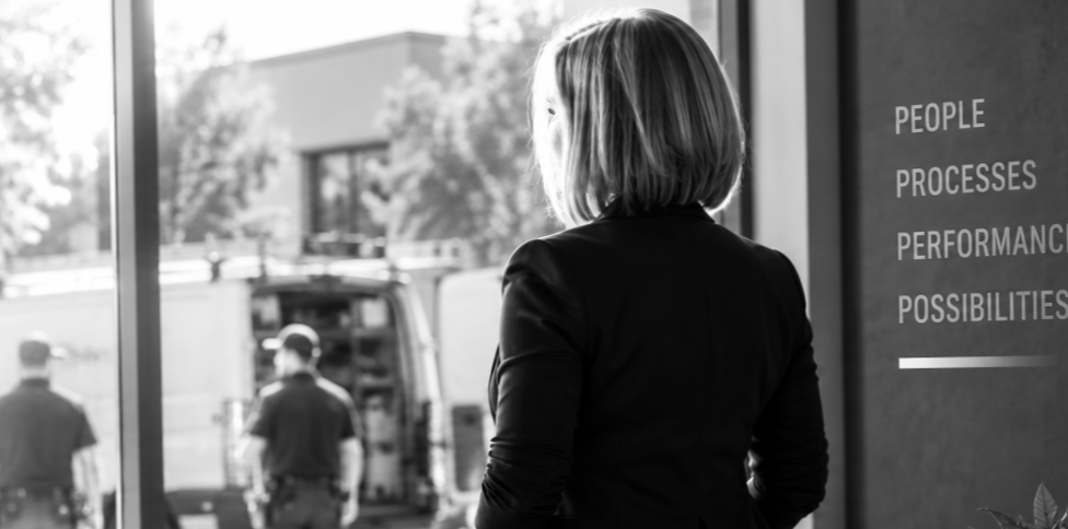 Woman looking out a window with police officers and a police vehicle outside at a building with words on the wall that read 'People, Processes, Performance, Possibilities'.