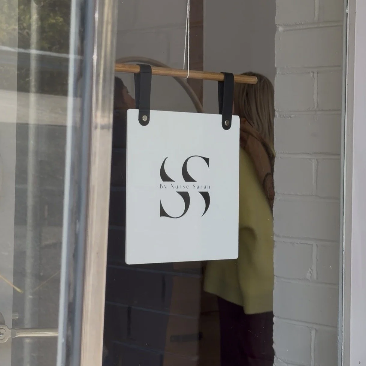 A storefront window with a white sign hanging from black straps that reads 'Be Nourse Sarah' with decorative initials 'SS'.