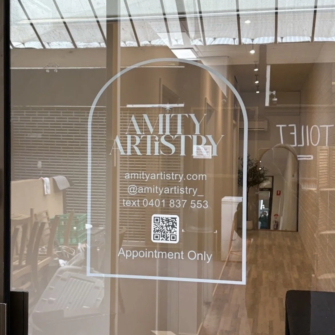 Glass door with the logo and contact information for Amity Artistry, including website, Instagram handle, phone number, and a QR code, with the note "Appointment Only."