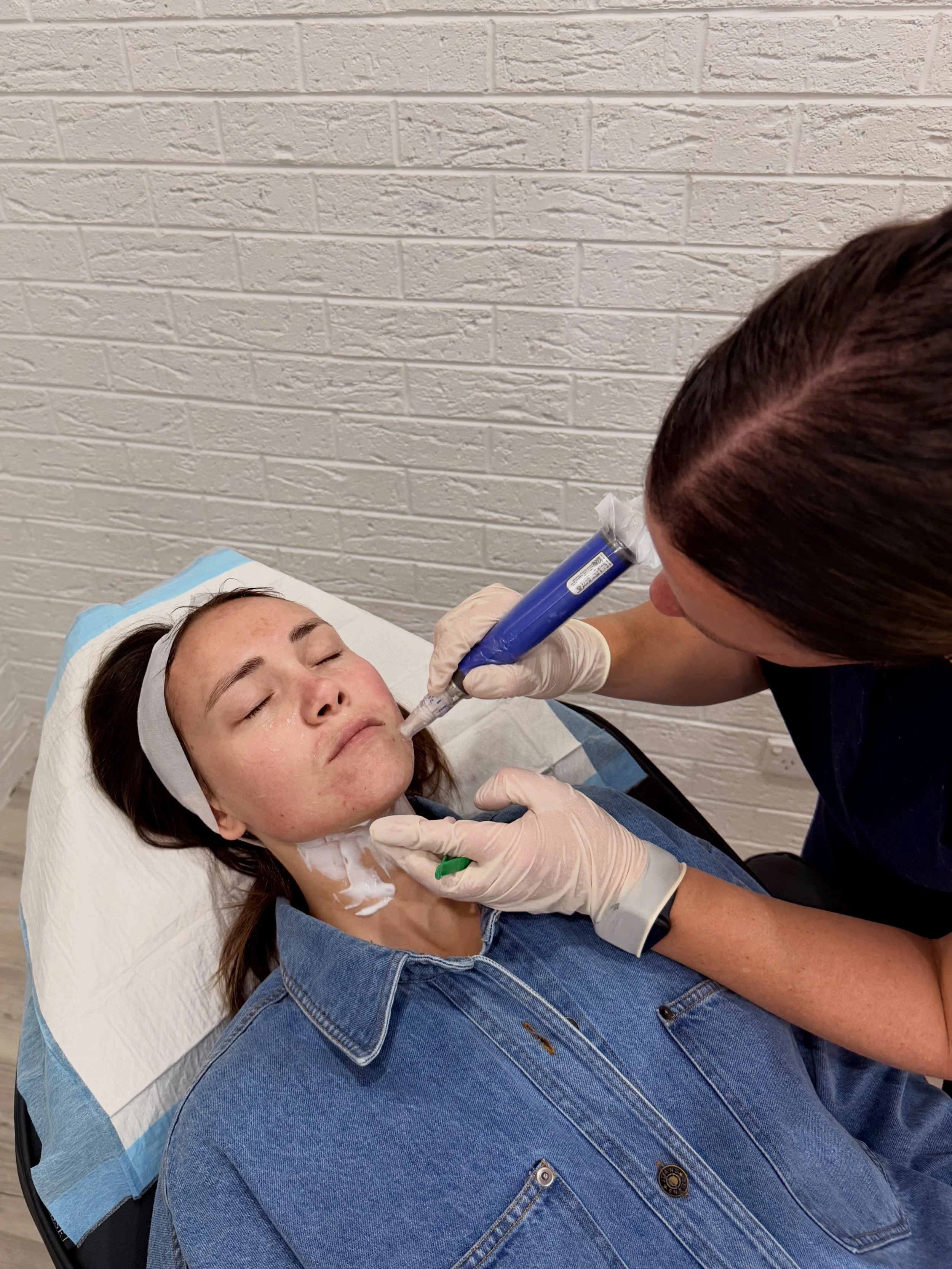 A woman lying on a medical bed receiving a cosmetic or dermatological treatment on her face from a professional practitioner using a handheld device.