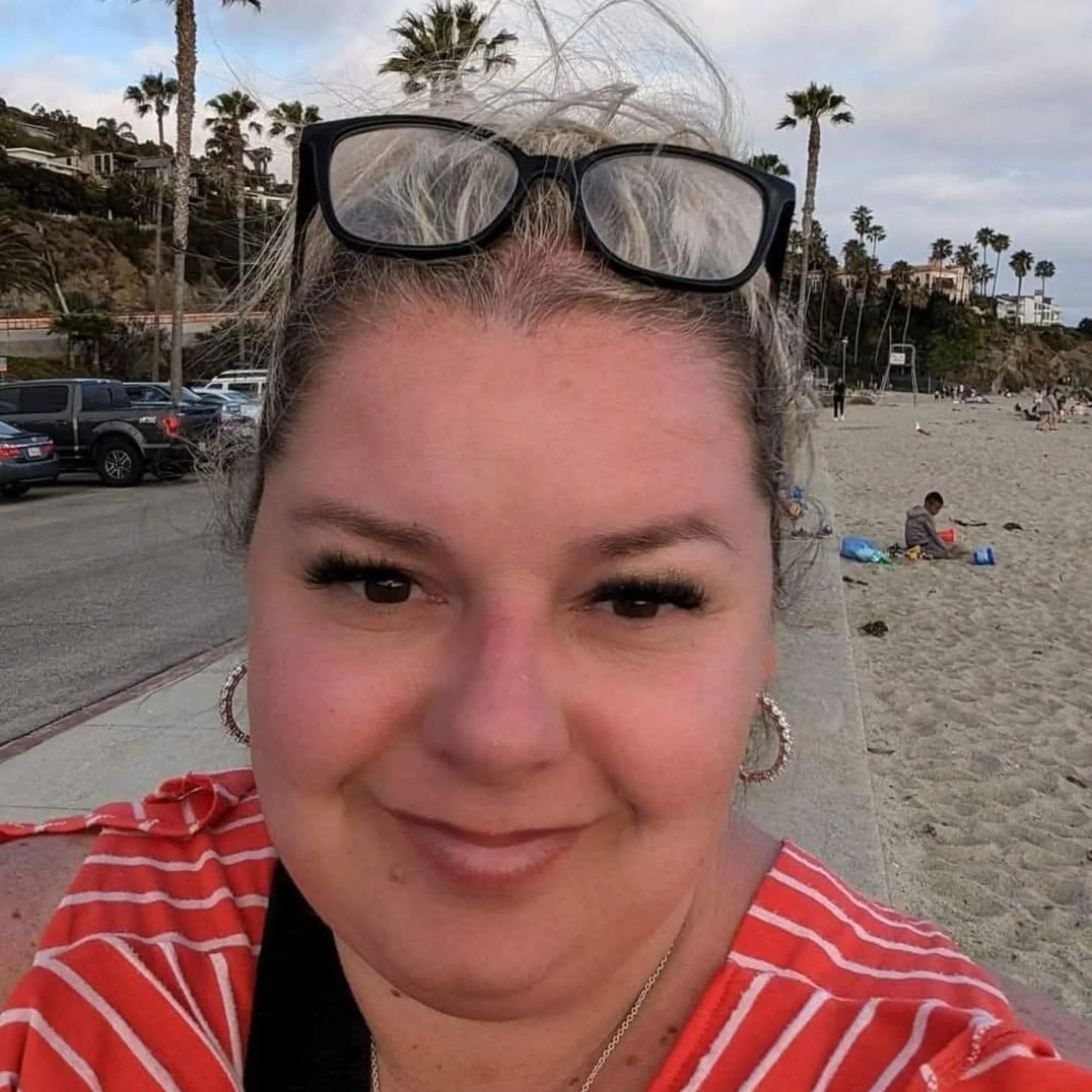 Close-up of a smiling woman with blonde hair, wearing black-rimmed glasses on her head, red and white striped shirt, at the beach with palm trees and houses in the background.