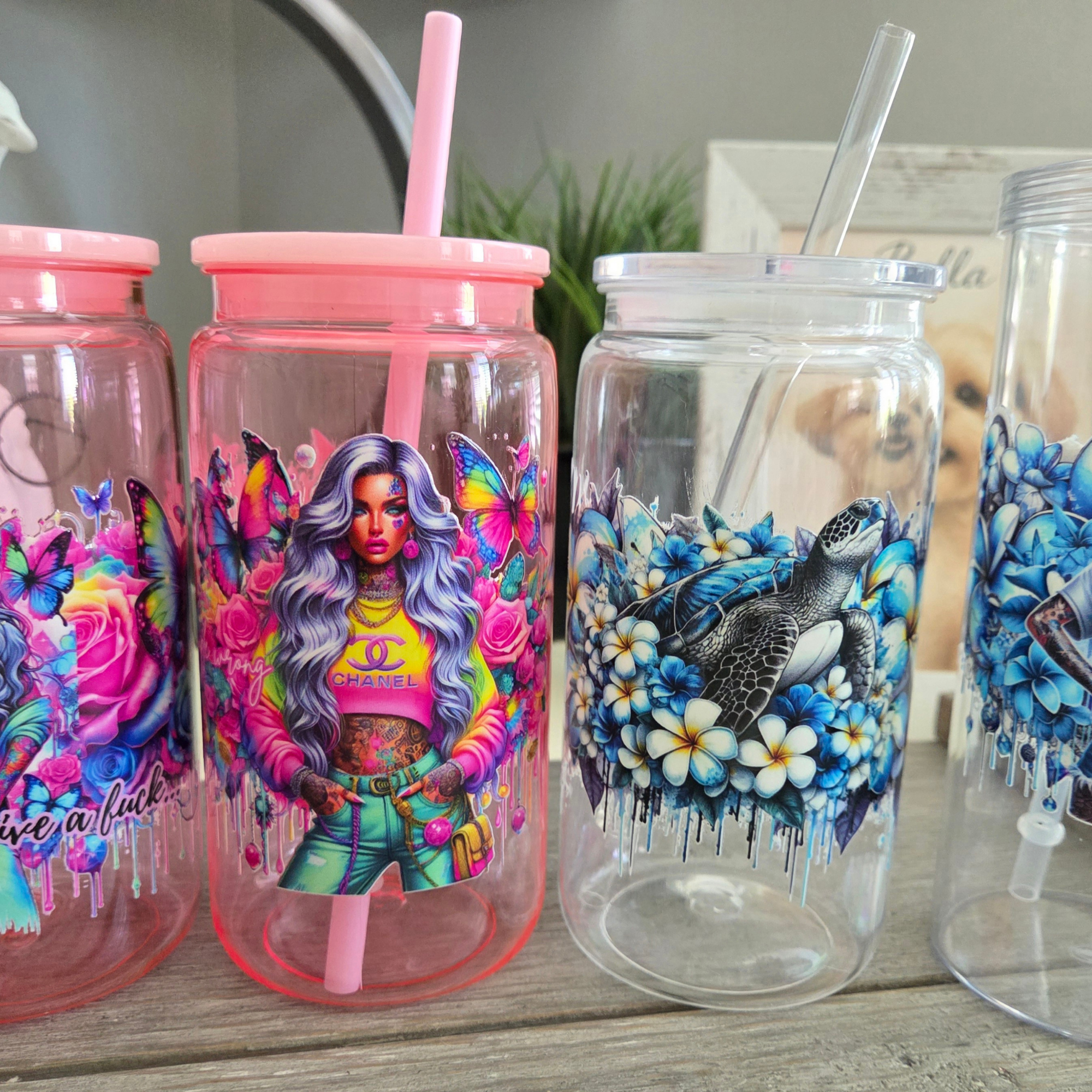 Clear plastic cups with colorful dragon and butterfly designs, some with pink and clear straws, arranged on a wooden surface with a plant and kitchen background.
