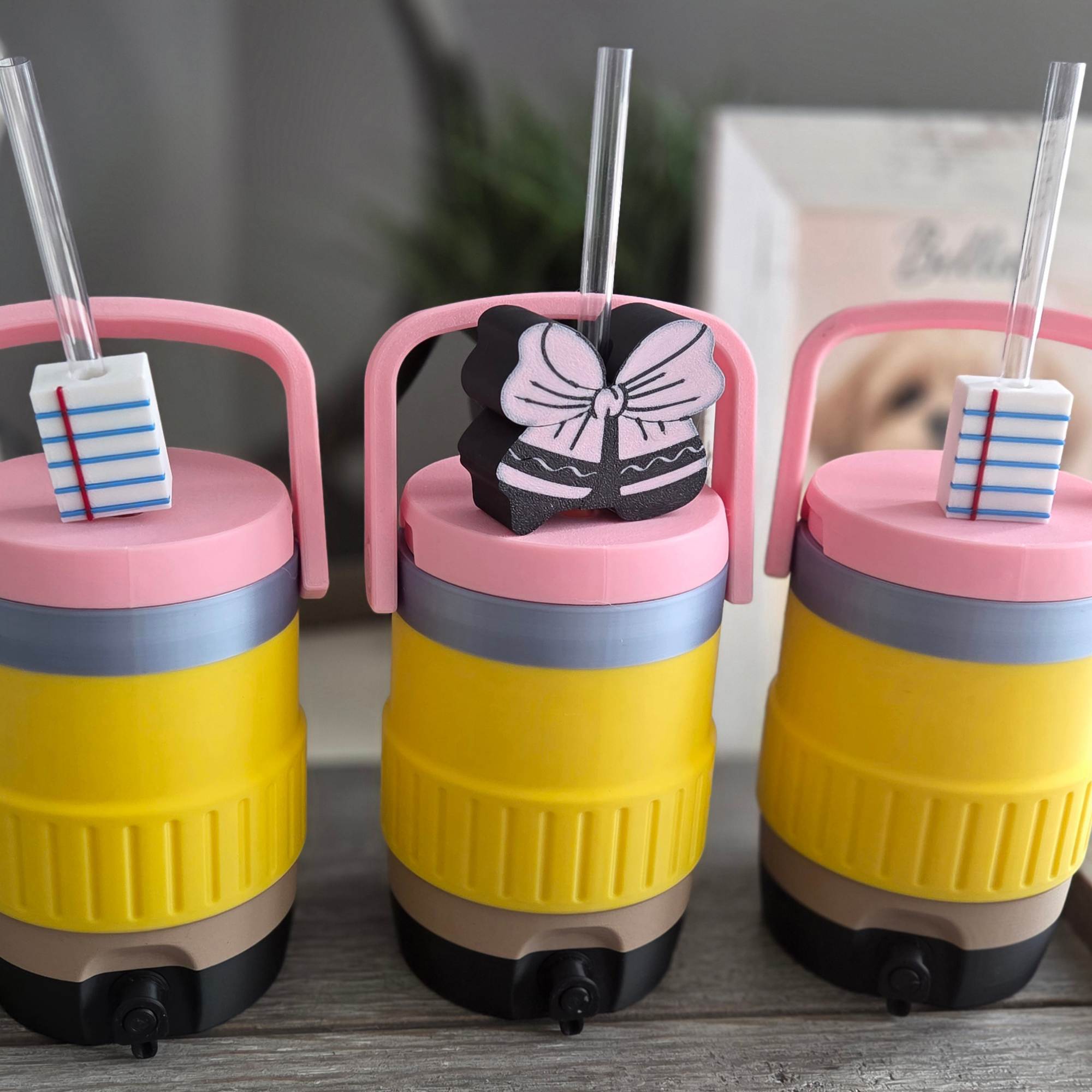 Close-up of four colorful glittery toy drink containers with pink, black, and yellow bases, featuring pink chairs with cut-out backrests, small wheels at the bottom, and clear straws, displayed on a wooden surface with a blurred background.