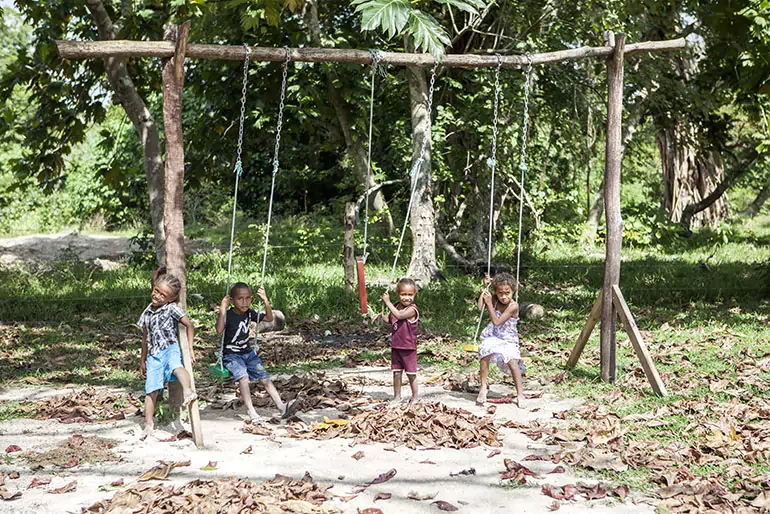 Four children playing on swings in a park surrounded by trees and dry leaves.