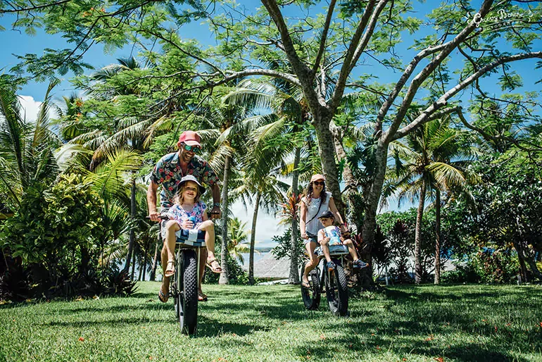A family of four riding bikes through a lush, tropical park with green grass, palm trees, and a clear blue sky.