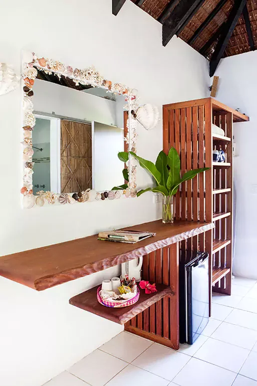 Interior view of a room with a wooden shelf, a mirror decorated with shells, a large green plant in a glass vase, and a small refrigerator beneath a wooden counter with a pink basket and magazines.