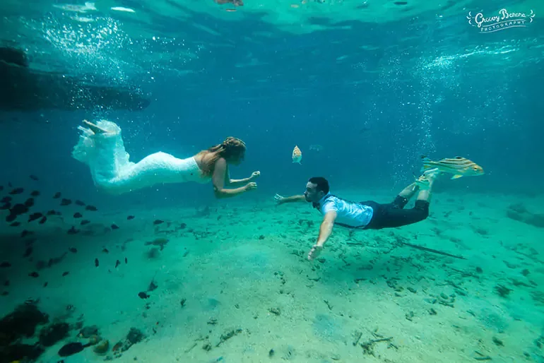 A man and woman are underwater, swimming towards each other, with the woman dressed as a mermaid and the man in casual clothing, surrounded by fish and underwater scenery.