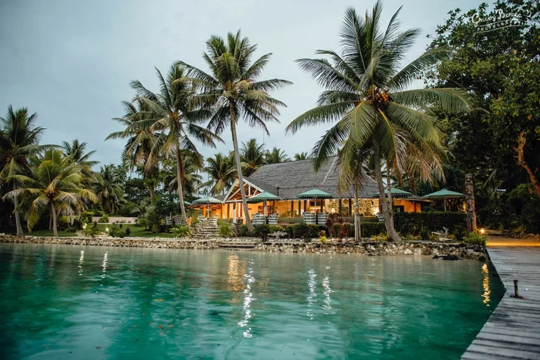 Tropical beachfront with palm trees, a house with a porch, and calm water in the foreground at sunset or dusk.