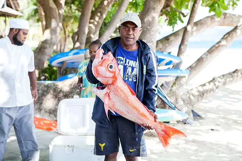 Man holding large red fish on the beach with kayaks and trees in the background.