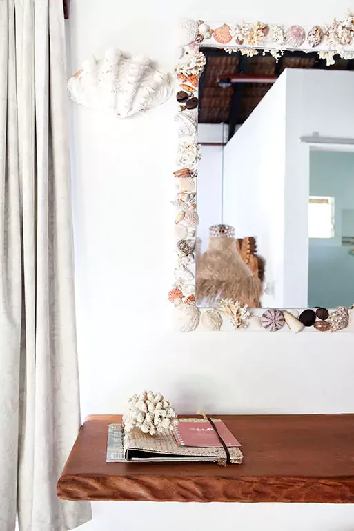 A wooden table with a lace flower arrangement on top, in front of a white wall with a mirror decorated with seashells; a curtain partially covers the left side of the image.