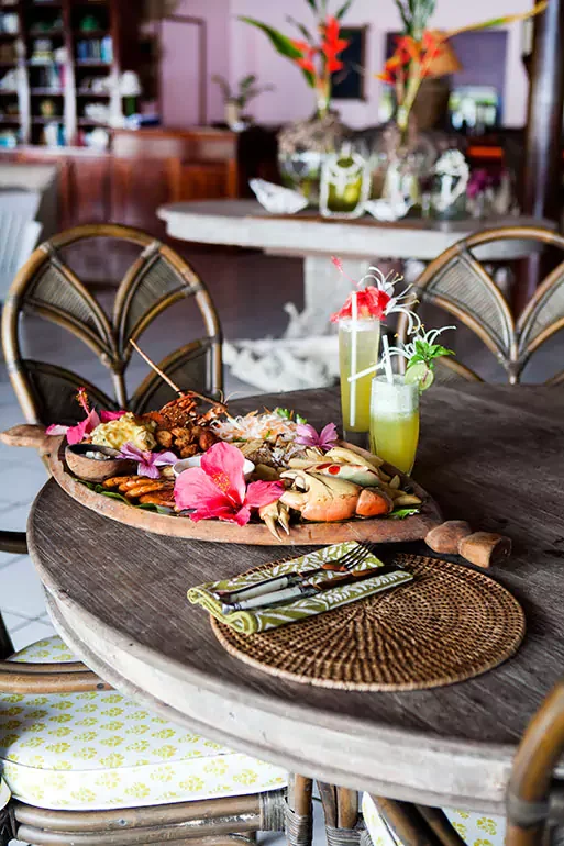 A wooden dining table set with drinks, a seafood platter with garnishes, and place settings in a tropical restaurant.
