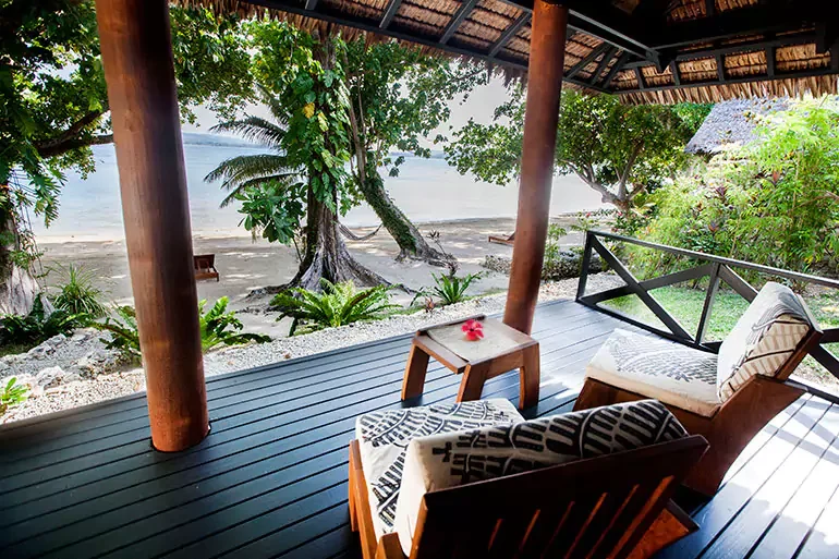Covered porch overlooking a sandy beach and ocean with tropical trees, plants, and seating with cushions.