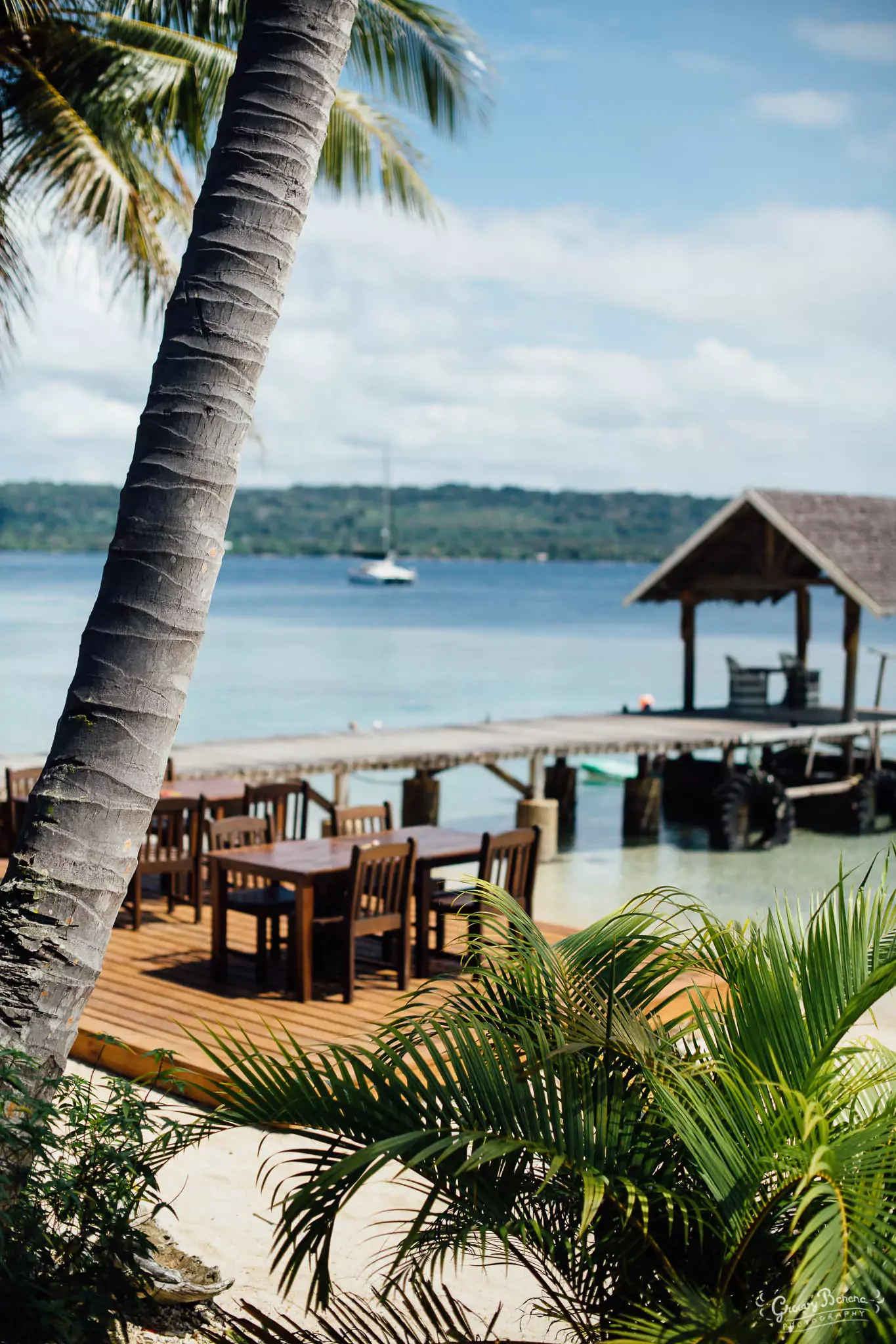 A tropical scene with a palm tree trunk in the foreground, a sandy beach area with wooden tables and chairs on a deck, water with a sailboat in the distance, and a thatched-roof hut by the water's edge.
