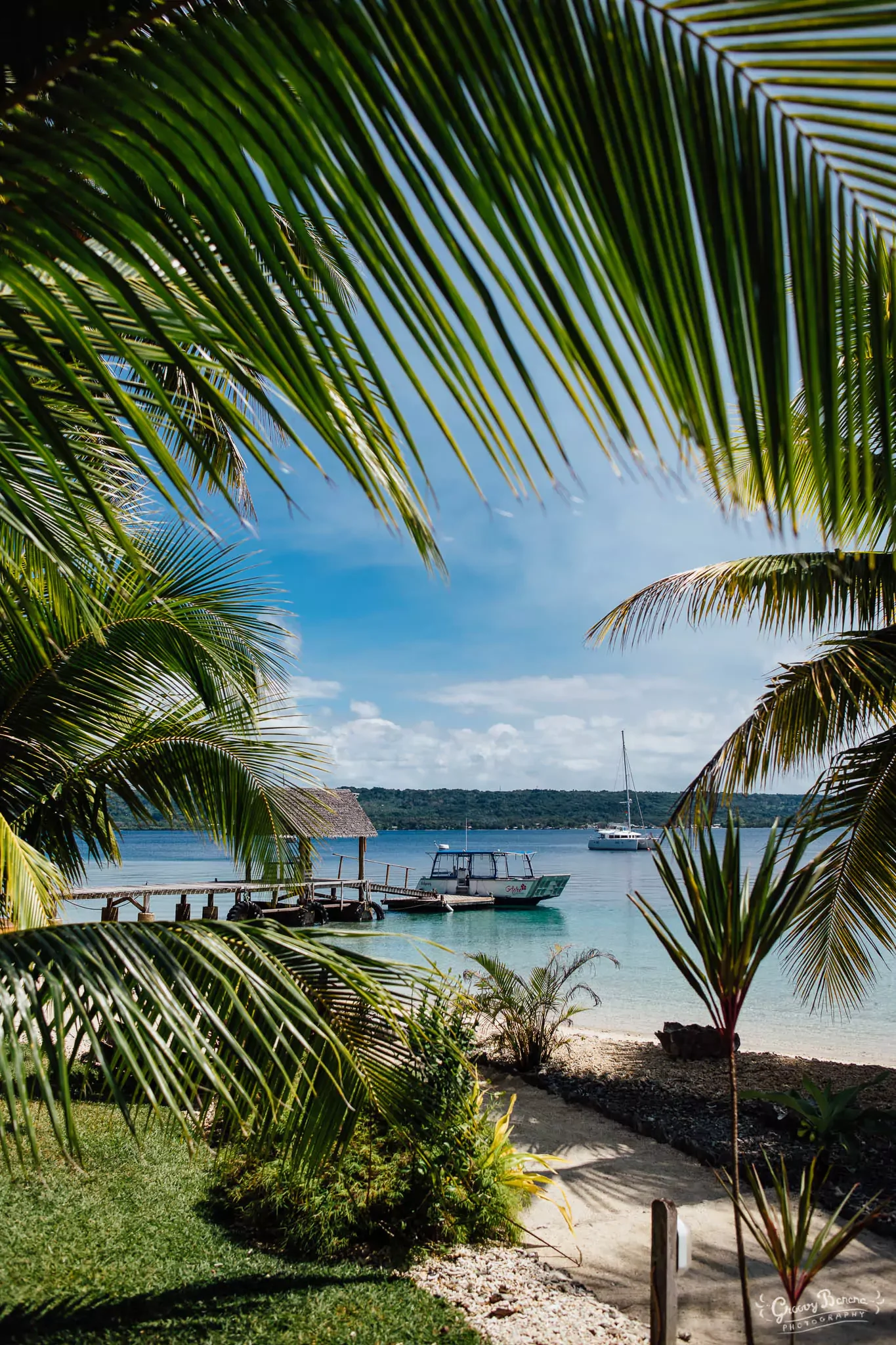 Tropical beach scene with palm trees framing view of docked boats on calm water, small hut, and distant hills under partly cloudy sky.