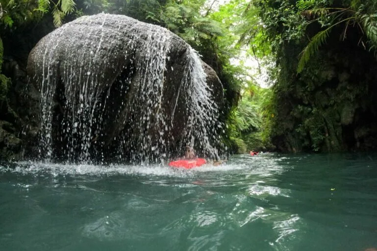 A small waterfall flowing over a large rock into a river surrounded by lush greenery.