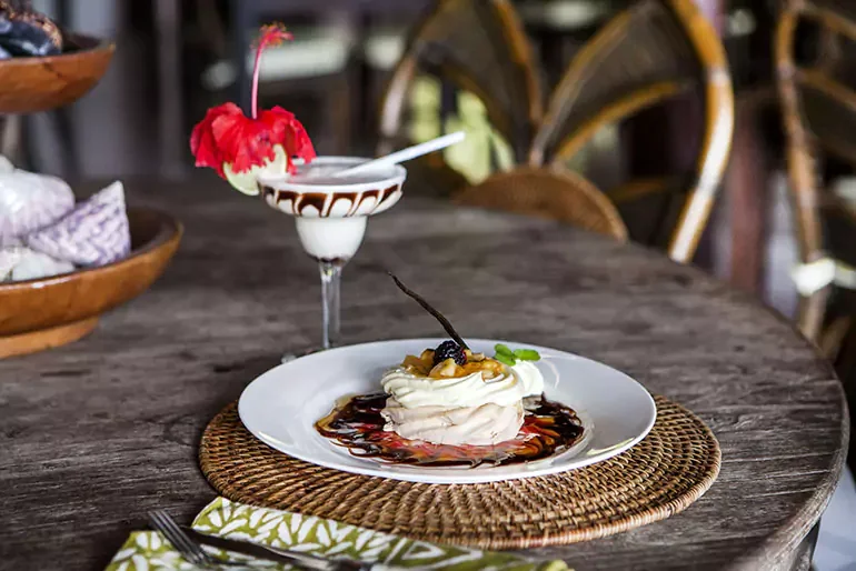 A dessert plate with a meringue topped with fruit and sauce, and a cocktail with a cherry and flower garnish, on a woven placemat on a rustic wooden table.