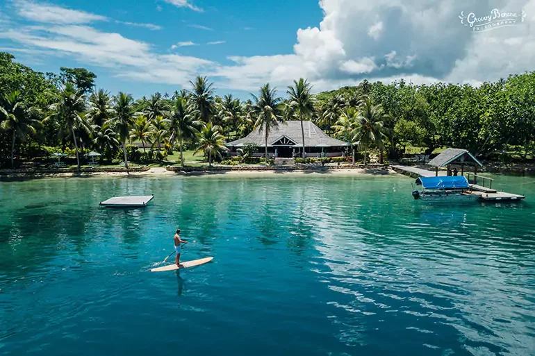 Person paddleboarding on clear blue water near a tropical shoreline with palm trees and a building with a thatched roof