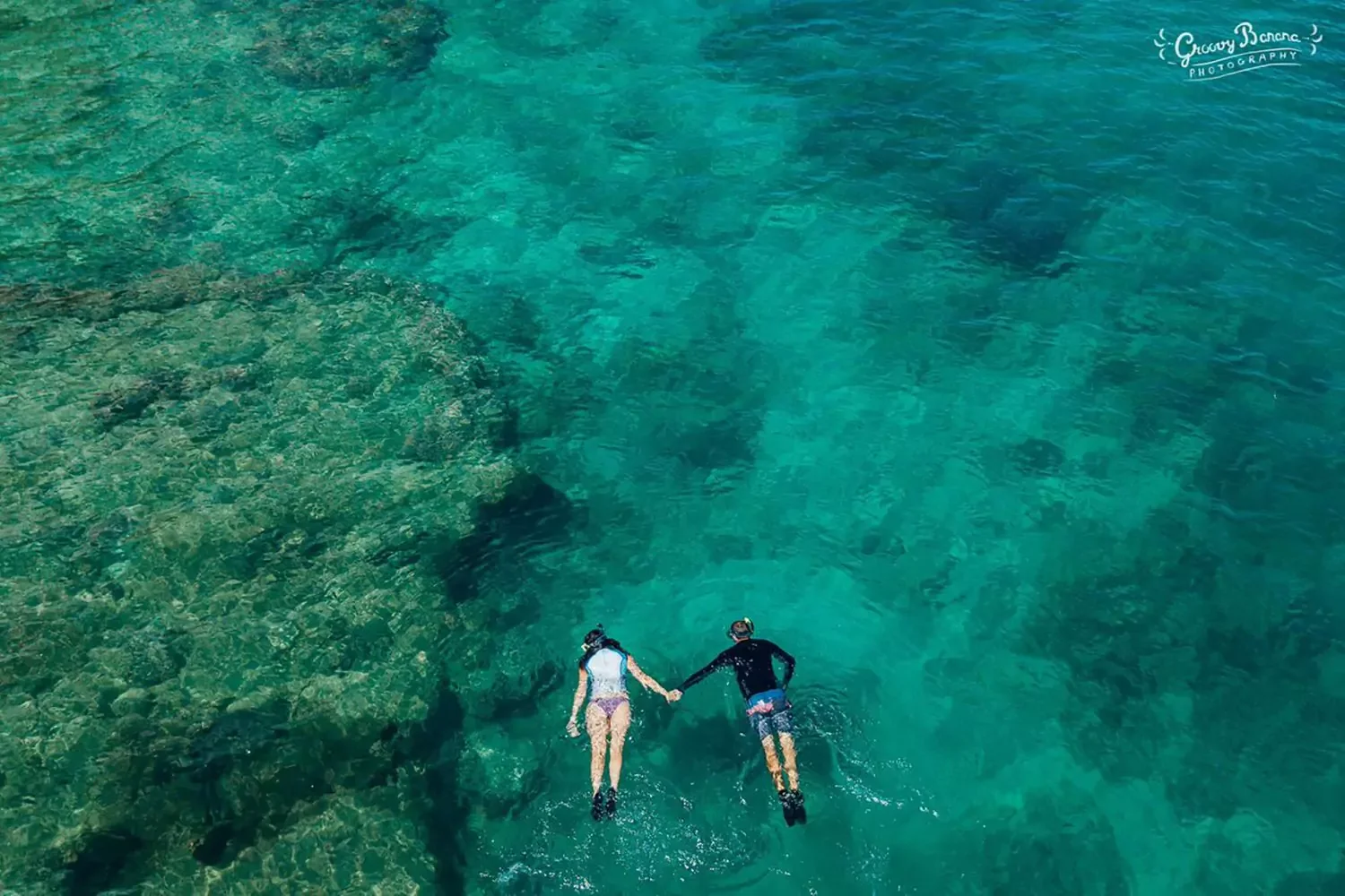 Two people in wetsuits and helmets holding hands and floating in clear blue water.