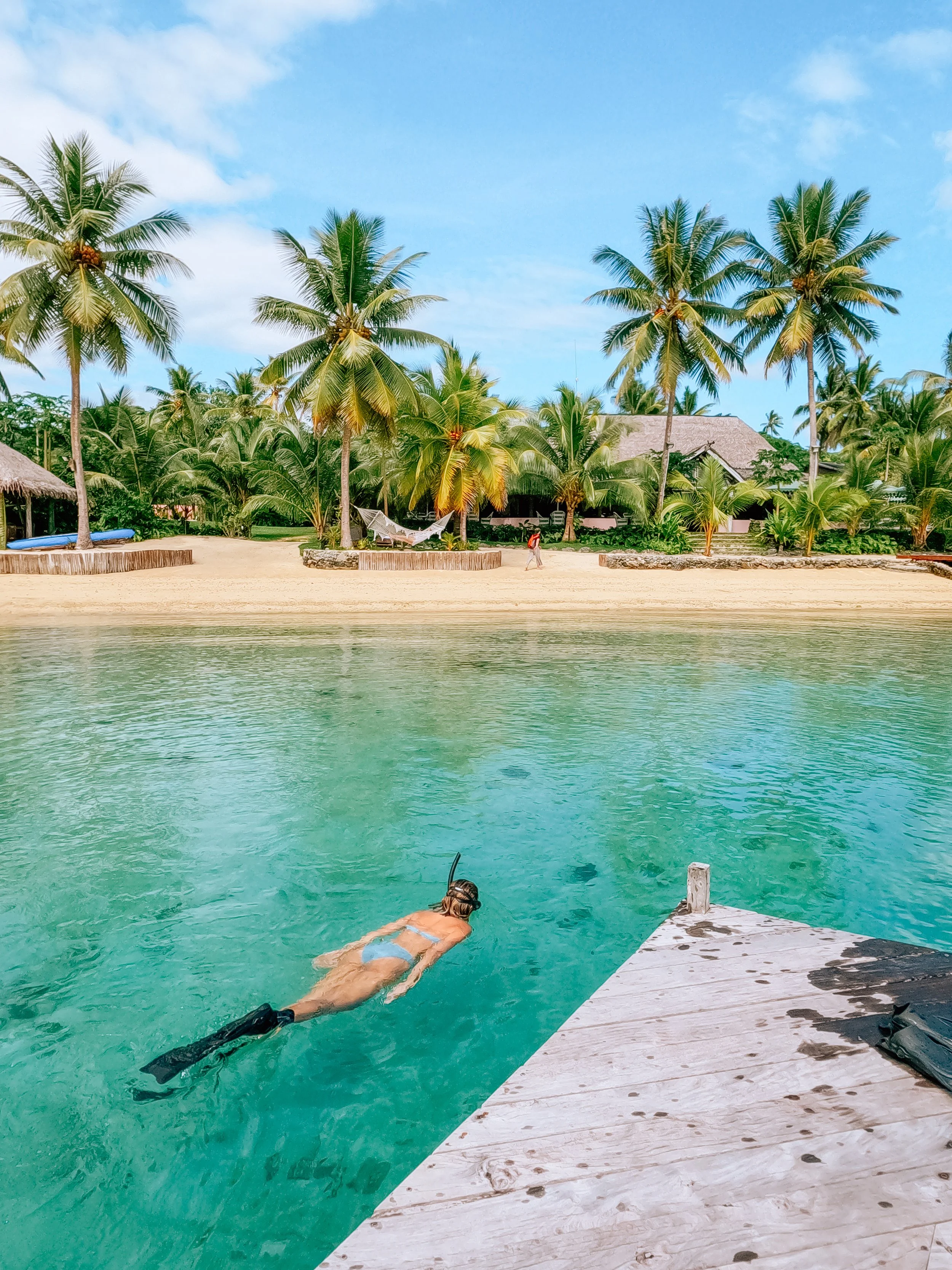 A person swimming in a clear blue-green body of water near a wooden dock, with a tropical backdrop of palm trees and thatched-roof buildings under a partly cloudy sky.
