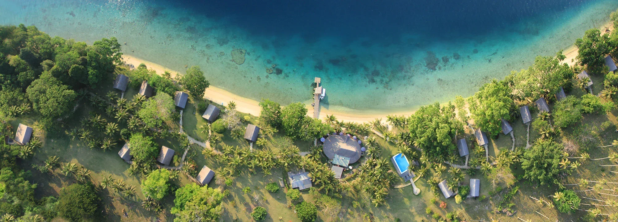 Aerial view of a tropical beach with clear blue water, white sand, surrounded by lush green trees and several small bungalows, some with thatched roofs, along the shoreline.