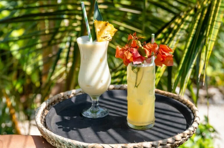 Two tropical cocktails on a black tray outdoors, with greenery in the background. One drink is a creamy white smoothie with a pineapple slice and a green straw. The other is a yellow drink with orange flowers and a white straw.
