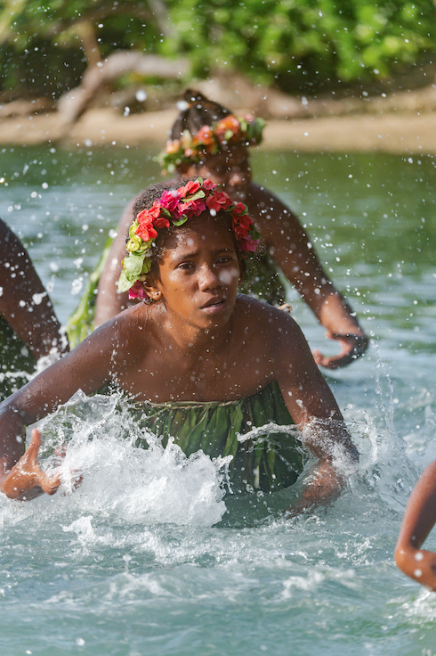 Young woman in green dress and floral crown in a river, participating in a traditional ceremony or celebration.