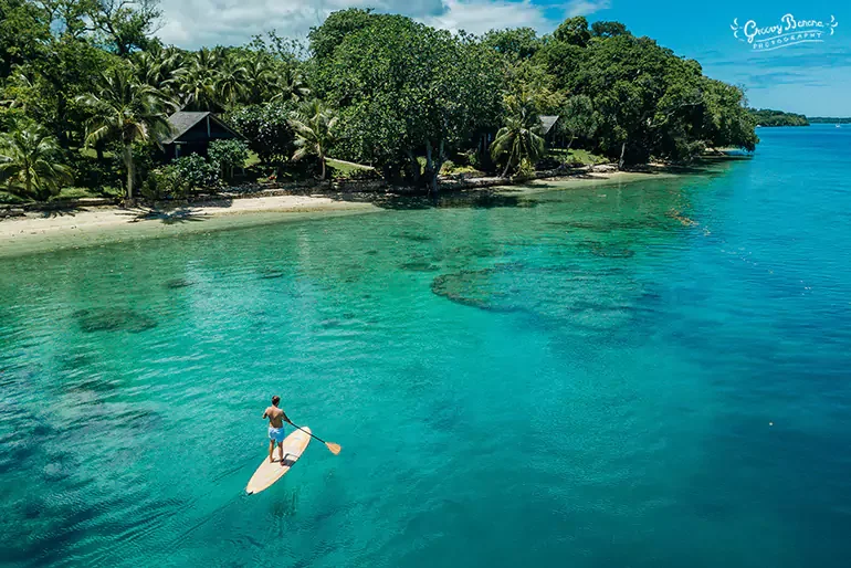 A person paddleboarding on clear turquoise water near a tropical shoreline with trees and small huts.