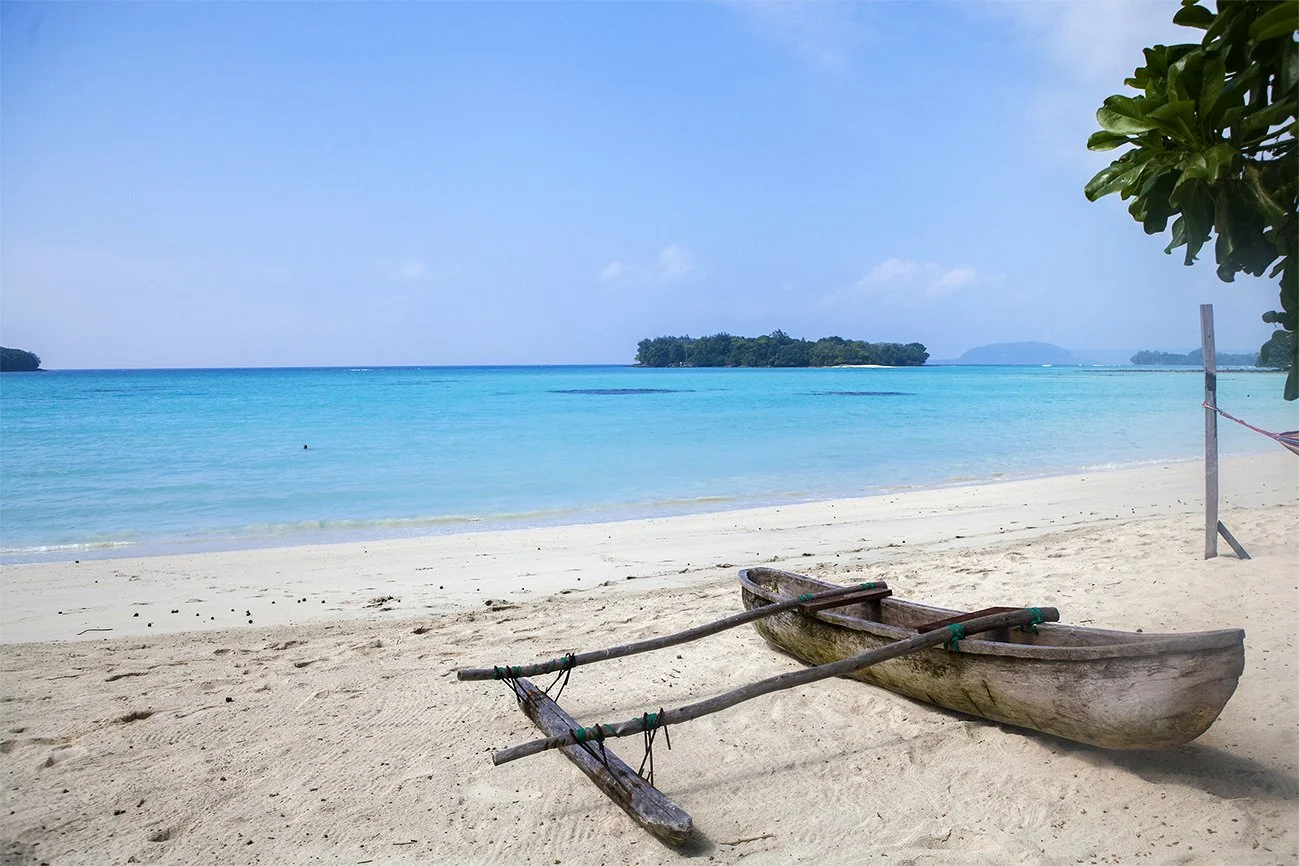 A wooden boat resting on a sandy beach near the turquoise ocean with small islands in the distance and a clear blue sky.