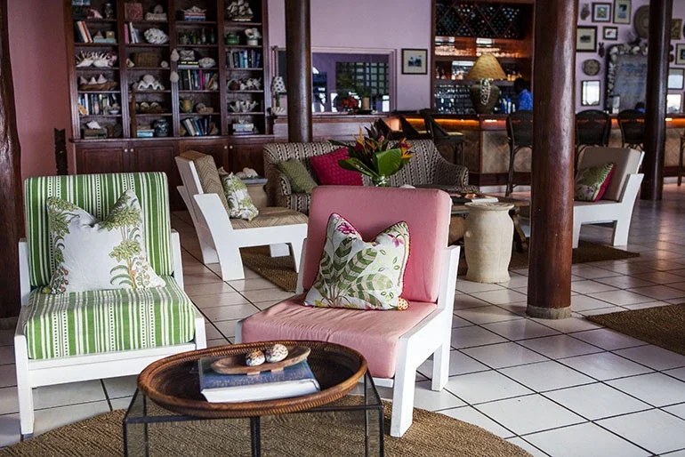 Cozy lobby area with colorful armchairs, pillows, and a round coffee table with shells and a book, set on tiled flooring with a bookshelf and decorative items in the background.