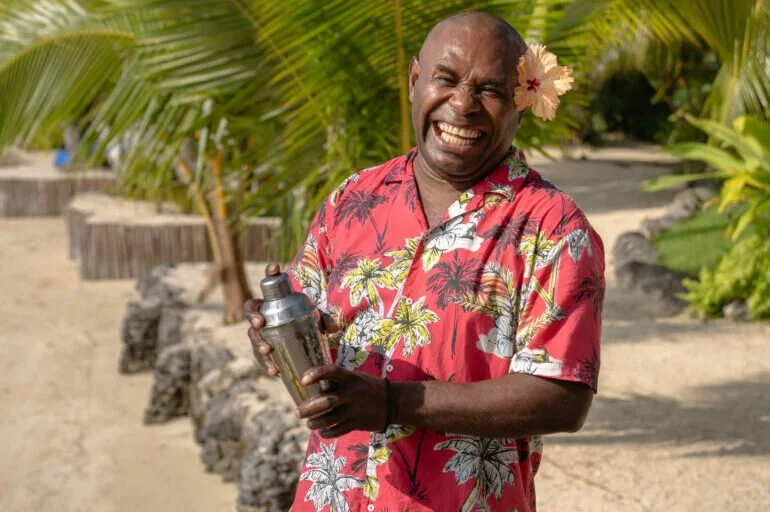 A smiling man with a flower behind his ear, wearing a red Hawaiian shirt with a palm tree pattern, holding a cocktail shaker on a sunny tropical beach surrounded by palm trees.