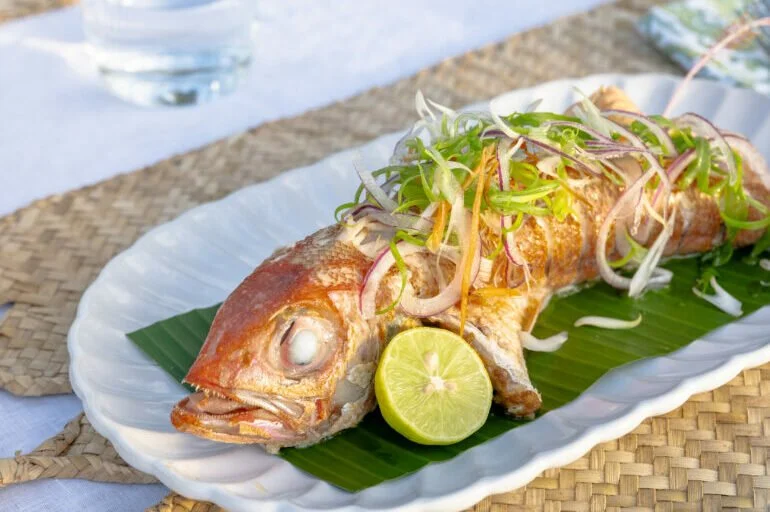 Whole fried fish topped with sliced red onions and green herbs, served with a lemon half on a banana leaf on a white platter.