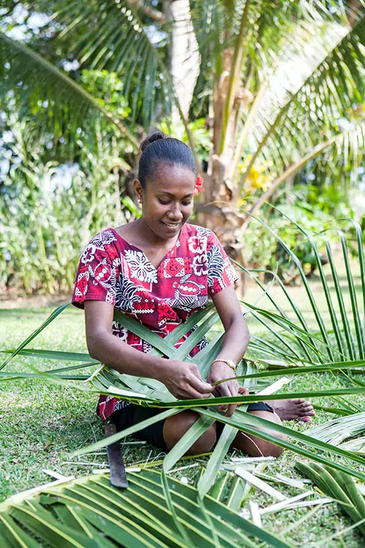A woman in a colorful floral top sits on the grass, weaving palm leaves in a lush, tropical setting with palm trees in the background.
