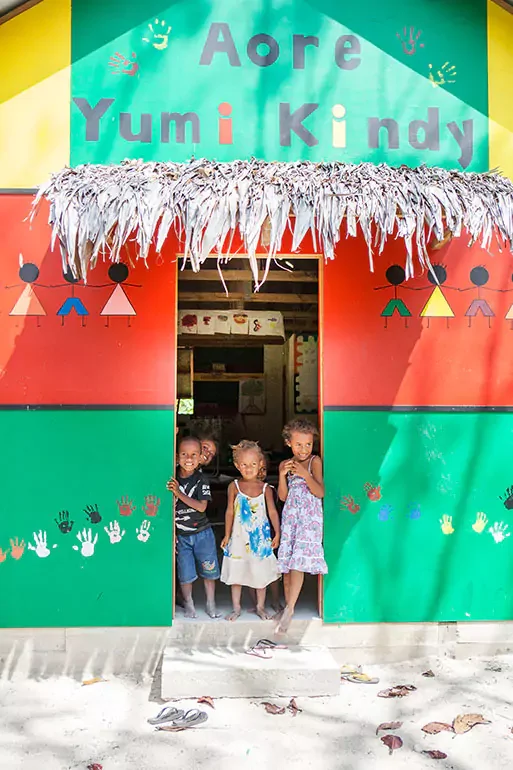 Three children standing in the doorway of a colorful building with a thatched roof. The building has painted handprints and symbols, with the words 'Aore Yumi Kindy' on the sign above. The children are smiling and appear happy.