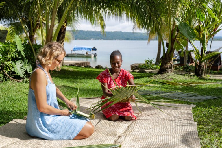 Two women sitting on a mat under palm trees by a lake, making traditional crafts with large palm leaves, with a boat on the water in the background.