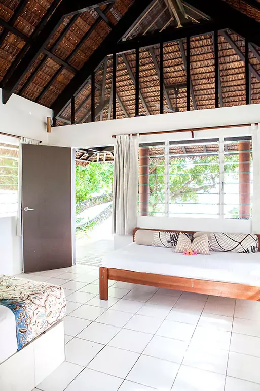 Interior of a bright room with a wooden bed, patterned pillows, white curtains, and large windows overlooking greenery. The ceiling is thatched with exposed wooden beams.