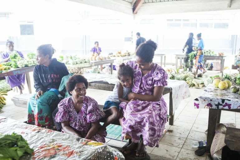 Group of women and children in a bright market or community space with tables of produce in the background.