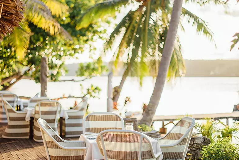 Outdoor restaurant with tables and chairs under palm trees near water, sunny day