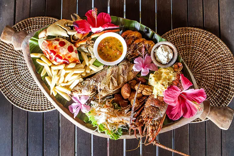 Seafood platter on a wooden table decorated with pink hibiscus flowers, including lobster, fish, shrimp, fries, sauces, and fresh herbs.