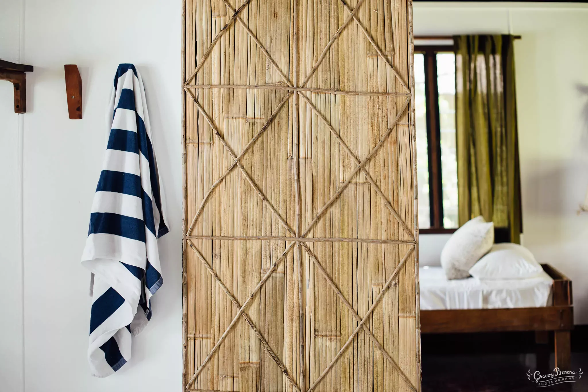 A striped towel hanging on a wooden hook against a white wall, with a bamboo room divider partially visible and a bedroom with green curtains and a bed in the background.