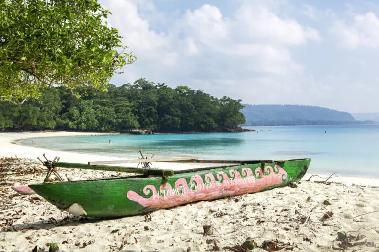 Green and pink painted boat resting on a sandy beach with clear turquoise water and lush green trees in the background.