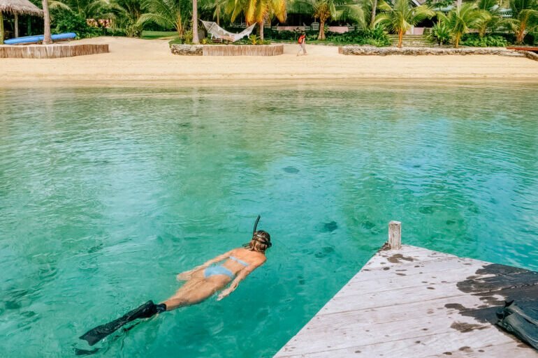 A person swimming in clear turquoise water near a wooden dock with a beach and palm trees in the background.
